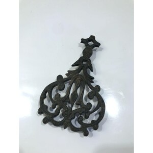 May include: Dark brown metal trivet with an intricate, openwork design. The trivet has a central floral motif and scrolled edges. The top has a cross-like shape. This vintage-style trivet is designed to protect surfaces from hot cookware.