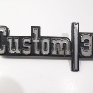 May include: A vintage car emblem featuring the word "Custom" in silver lettering on a black base, with the number "30" to the right. The emblem has a rectangular shape with a vertical bar.