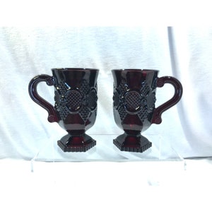 Puede incluir: Dos tazas de vidrio rojo con un diseño en relieve y una base con pie. Las tazas tienen un asa y están hechas de vidrio rojo rubí.