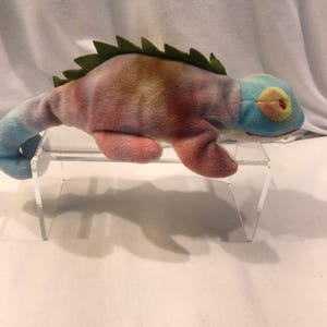 Peut inclure: Une peluche caméléon avec un motif tie-dye aux teintes bleues, roses et orange. Ce jouet en peluche présente une texture douce, une crête en feutrine vert foncé sur le dos, une queue enroulée et une tête bleu clair avec des taches oculaires jaunes.