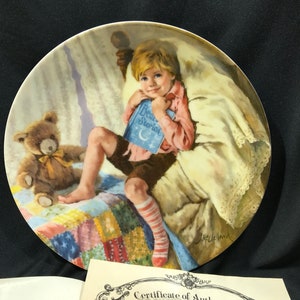 Könnte beinhalten: Ein Porzellan-Teller mit einer farbenfrohen Illustration eines jungen Jungen im Schlafanzug, der ein Buch mit dem Titel "Bedtime Stories" liest, während er mit einem Teddybär im Bett sitzt. Der Teller ist mit einer Patchwork-Decke verziert und trägt eine Signatur in der rechten unteren Ecke.