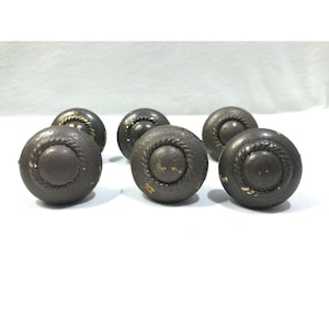 Pode incluir: Um conjunto de seis puxadores redondos de metal castanho escuro para armários. Cada puxador apresenta uma cúpula central circular lisa rodeada por uma borda elevada em forma de corda torcida. As superfícies metálicas mostram sinais de desgaste.