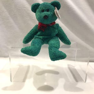 Peut inclure: Un ours en peluche vert d'eau avec une écharpe à carreaux rouge et noir. L'ours a des yeux en boutons noirs et un nez noir. Il est assis sur un présentoir en acrylique transparent. Une étiquette blanche est attachée à l'oreille de l'ours.