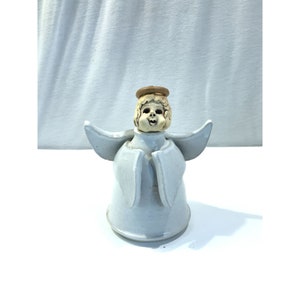 Puede incluir: Figura de ángel de cerámica con un esmalte azul claro. El ángel tiene una cara sonriente, un halo y alas extendidas. La figura está sobre una superficie blanca.