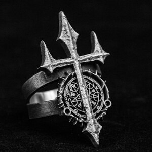 Op de afbeelding: Een zilveren ring met een gedetailleerd ontwerp van een kruis met een drietand aan de bovenkant en een cirkelvormig patroon met ingewikkelde details. De ring is gemaakt van metaal en heeft een textuur.