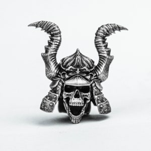 May include: A silver skull wearing a detailed samurai helmet with horns. The skull has an open mouth and visible teeth.