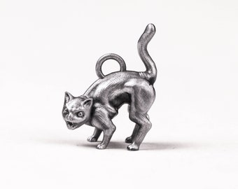 Puede incluir: Un amuleto de plata con forma de gato, con la espalda arqueada y la cola levantada, como si estuviera silbando. El gato tiene una cara detallada con dientes afilados y un pequeño bucle en la parte superior de la cabeza para sujetarlo a un collar o pulsera.