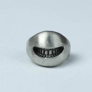 May include: A silver ring with a detailed, toothy grin design. The ring is smooth and has a brushed finish.