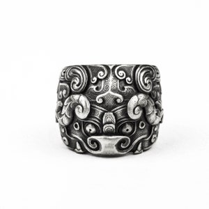 May include: A silver ring with an intricate design of a mythical creature's face. The design features swirling patterns and a detailed face with large eyes.