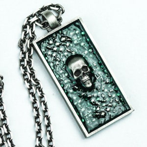 Könnte beinhalten: Eine silberne Kettenkette mit einem rechteckigen Anhänger, der einen silbernen Schädel zeigt, der von kleineren Schädeln auf einem grünen Harzgrund umgeben ist.