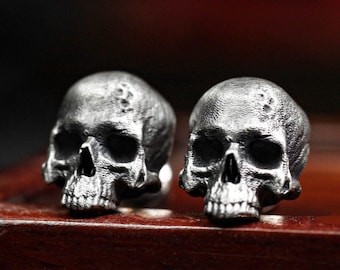 Può includere: Due gemelli a forma di teschio in argento, con superfici testurizzate, sono esposti su una superficie in legno scuro.