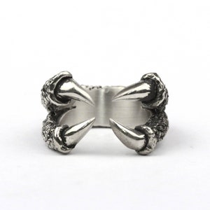 May include: A silver ring featuring a detailed design of two claws, each with a sharp point. The ring is made of metal and has a textured surface.