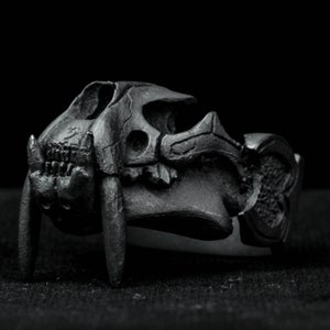 Puede incluir: Un anillo de metal negro con un diseño de calavera detallado. La calavera tiene dientes afilados y una superficie texturizada.
