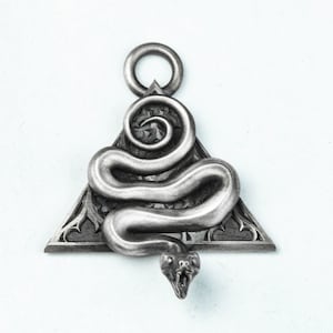 Può includere: Un ciondolo d'argento a forma di triangolo con un serpente avvolto attorno. La testa del serpente è rivolta in avanti con la bocca aperta. Il ciondolo ha un anello nella parte superiore per attaccare una catena.