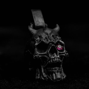 May include: A dark grey skull pendant with horns and a red gemstone eye. The pendant has a loop at the top for a chain and is set against a black background. The skull has detailed features and a textured surface.