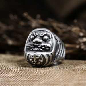 Könnte beinhalten: Ein silberner Ring mit einem detaillierten Design eines japanischen Wächterlöwen, bekannt als Shisa, mit einer strukturierten Oberfläche.
