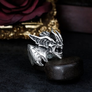 Puede incluir: Un anillo de plata con un diseño detallado y texturizado de una criatura con una boca grande abierta. El anillo está sobre una superficie gris oscuro.