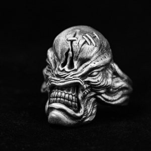 May include: A silver ring with a detailed, textured design of a creature's face. The creature has a large, open mouth with sharp teeth and a furrowed brow.