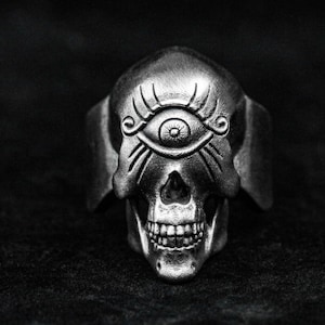 Può includere: Un anello a forma di teschio in argento con un designo di occhio onniveggente. L'occhio è circondato da dettagli intricati e il teschio ha una mascella dettagliata.