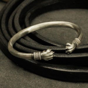 Op de afbeelding: Zilverkleurige manchetarmband met klauwontwerp. De armband heeft twee klauwvormige uiteinden, elk met ingewikkelde details. De klauwen zijn verbonden door een gladde, gebogen band. De armband wordt gepresenteerd op een donker, getextureerd oppervlak.