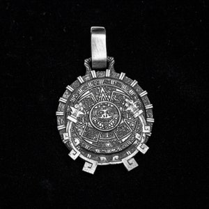 May include: Silver pendant featuring a detailed depiction of the Aztec calendar, also known as the Sun Stone. The pendant is circular with a raised rim and intricate carvings.