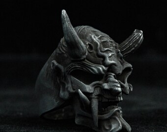 May include: A silver ring featuring a detailed, textured design of a demon mask. The mask has sharp horns, a fierce expression, and a set of fangs.