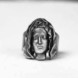 Könnte beinhalten: Ein silberner Ring mit einer detaillierten Skulptur eines Frauenkopfes, der eine Kapuze trägt.