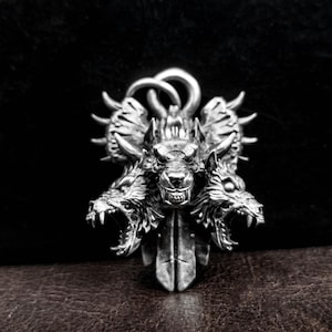 May include: A silver pendant featuring a three-headed wolf design. The wolf heads are snarling and have sharp teeth. The pendant is detailed with intricate carvings and a textured surface.