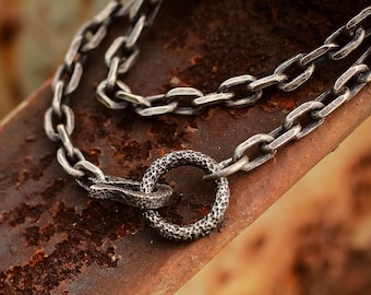 May include: Close-up of a silver chain necklace with a textured circular clasp. The chain links are rectangular and connected. The necklace is displayed on a rusty, brown surface, creating a contrast between the metal and the background.