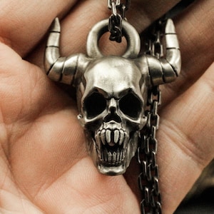 Può includere: Un ciondolo a forma di teschio in argento con corna, dettagliato con denti affilati e orbite vuote. Il ciondolo è attaccato a una catena d'argento.