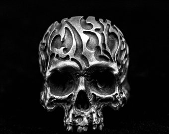 May include: A silver skull ring with intricate, detailed carvings on the skull. The skull has a tribal or gothic design.