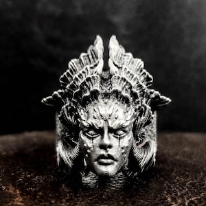 May include: A silver ring with a detailed design of a woman's face with horns and a crown. The ring is made of metal and has a detailed design.