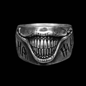 Puede incluir: Un anillo de plata con un diseño detallado de una boca sonriente con dientes afilados y la palabra "HAHA" a cada lado.