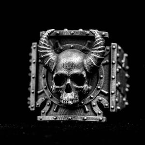 Può includere: Un anello dettagliato in scala di grigi con un teschio con corna ricurve e un design corazzato borchiato. Il teschio è centrato su un elemento a forma di scudo, con dettagli intricati e un'estetica gotica.