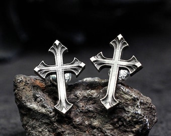 May include: A pair of silver cross earrings with intricate detailing. The earrings are on a dark gray rock surface.