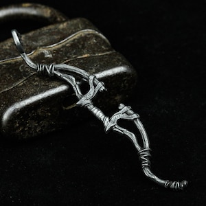 May include: A silver metal bow and arrow pendant, with a detailed design of a branch with leaves. The pendant is lying on a dark surface.