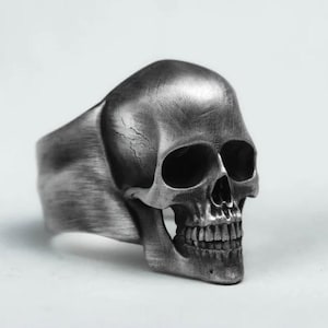 May include: A silver skull ring with detailed facial features. The ring has a wide band and a textured finish. The skull's eye sockets, nose cavity, and teeth are clearly defined, giving it a gothic or biker aesthetic.