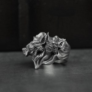 May include: A silver ring with two bat-like creature heads with fangs and wings. The heads are facing forward and the wings are spread out behind them.