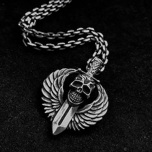 Może przedstawiać: Srebrny naszyjnik z zawieszką przedstawiającą czaszkę z skrzydłami i mieczem. Zawieszka jest przymocowana do srebrnego łańcuszka.