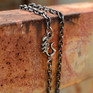 May include: Close-up of a silver chain necklace with a decorative clasp. The chain links are small and oval-shaped. The clasp has an ornate, stylized design. The necklace is displayed against a rusty, textured background.