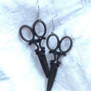 May include: A pair of bronze-coloured scissor earrings with silver hooks. The earrings are in the shape of a pair of scissors with a decorative design.