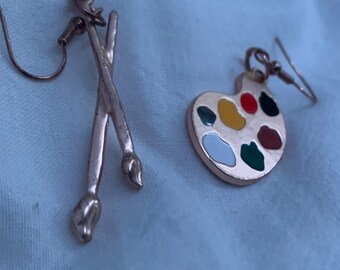 May include: A pair of gold-tone earrings. One earring is a paint palette with colorful enamel paint. The other earring is a pair of crossed paintbrushes.