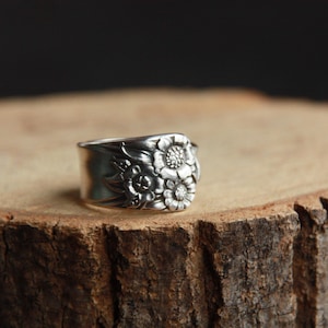 Può includere: Un anello d'argento con un design floreale, appoggiato su una superficie di legno. L'anello presenta dettagliati motivi di fiori e foglie. L'anello è largo circa 2 cm.