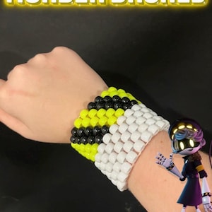 Peut inclure: Un bracelet de perles noir, jaune et blanc, avec le texte "MURDER DRONES" en jaune brillant. Un personnage de dessin animé est également visible.