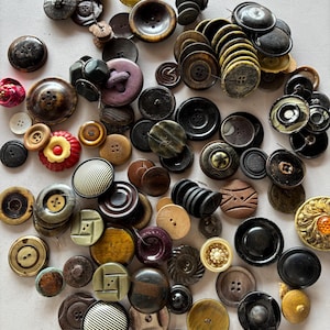 May include: A collection of various vintage buttons in shades of brown, black, and cream. The buttons are made of different materials, including wood, plastic, and metal, and feature a variety of shapes and sizes. Some have intricate designs.