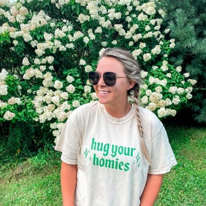 May include: A woman wearing sunglasses and a cream colored t-shirt with the text "hug your homies" in green lettering. She is standing in front of a bush with white flowers.