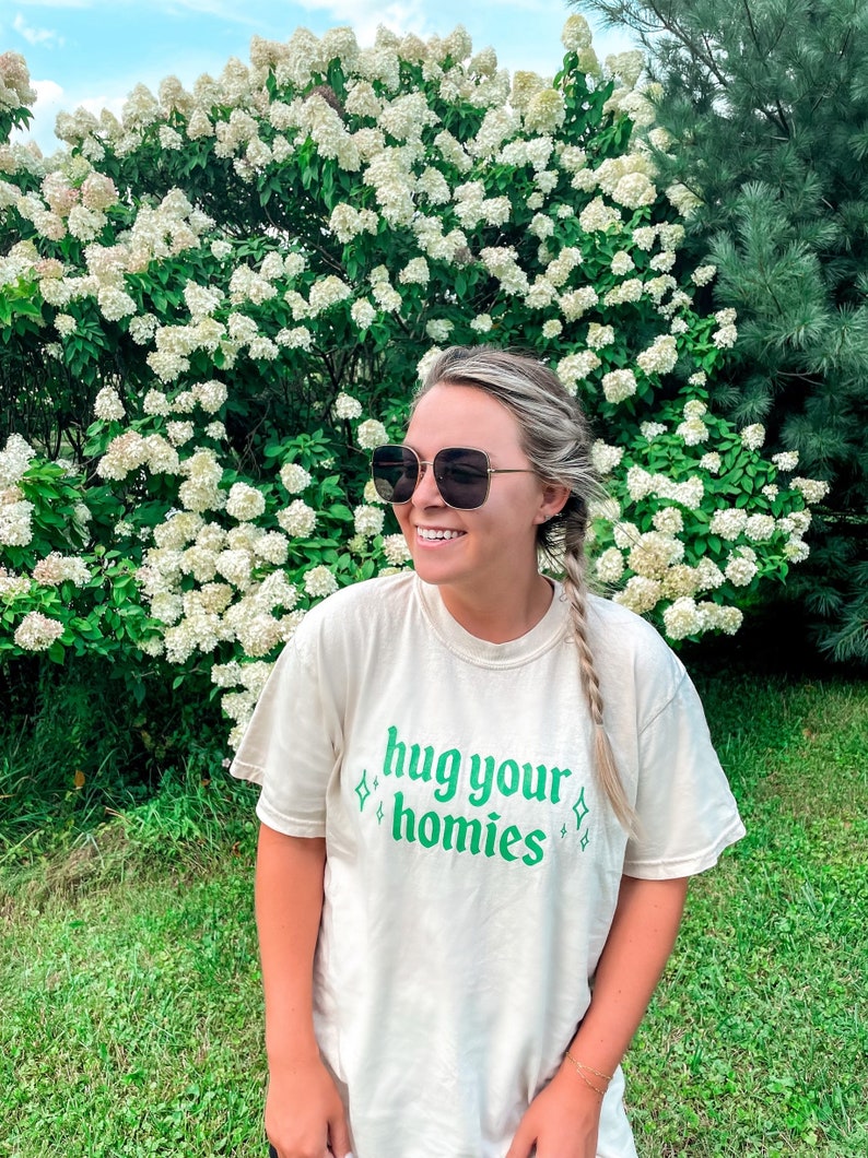May include: A woman wearing sunglasses and a cream colored t-shirt with the text "hug your homies" in green lettering. She is standing in front of a bush with white flowers.