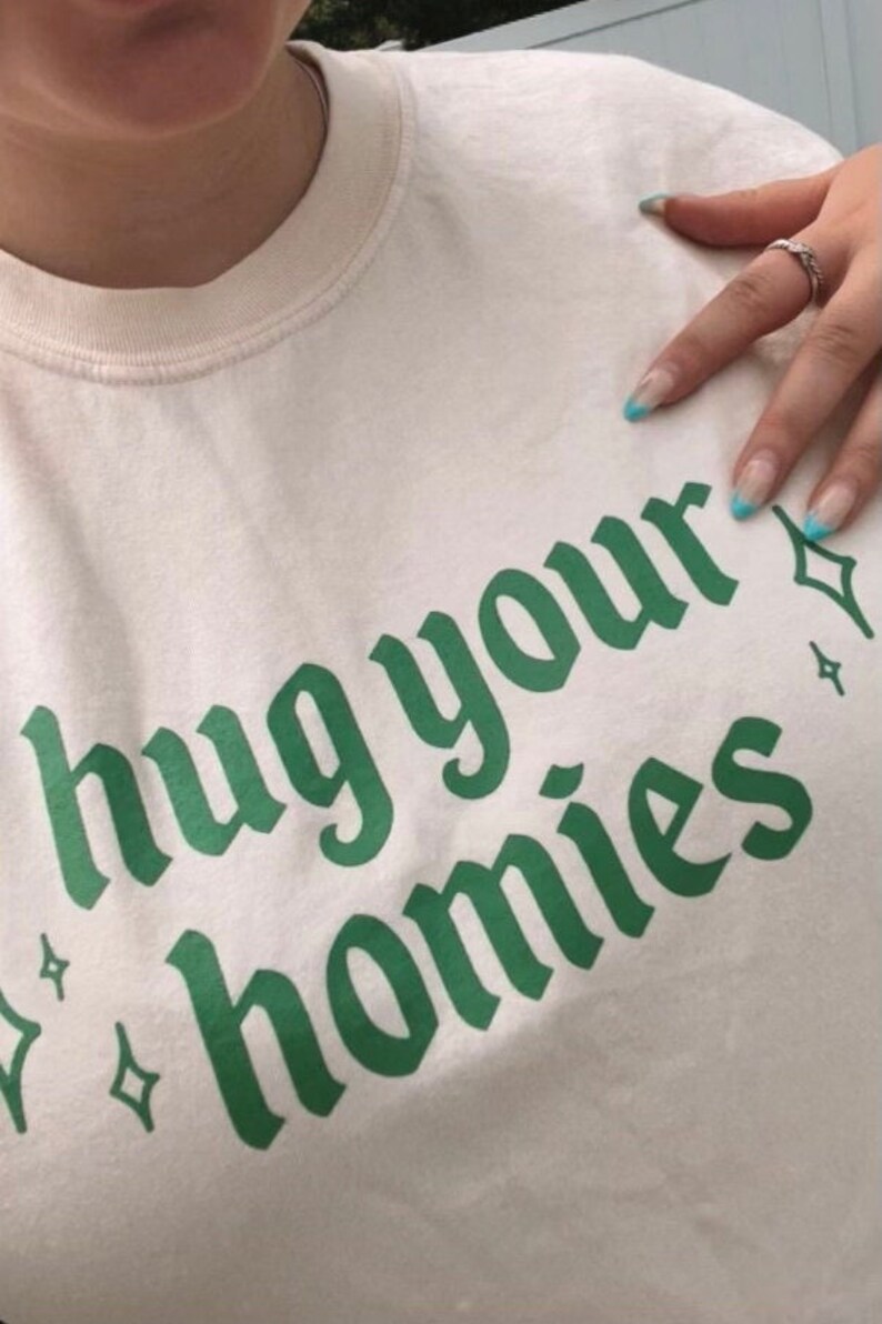 May include: A white t-shirt with the text "hug your homies" printed in green letters. The letters are in a gothic font style.