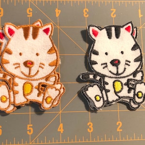 May include: Two embroidered patches of cartoon tiger cubs. One tiger cub is white with brown stripes and accents, the other is white with black stripes and accents. Both have red ears, yellow bellies, and are smiling.