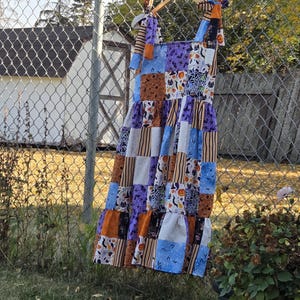 May include: A patchwork dress with a Halloween theme, hanging on a wooden hanger. The dress features fabrics in orange, purple, blue, and white, with patterns including pumpkins, bats, and spiderwebs. The dress has tie straps and a tiered skirt.